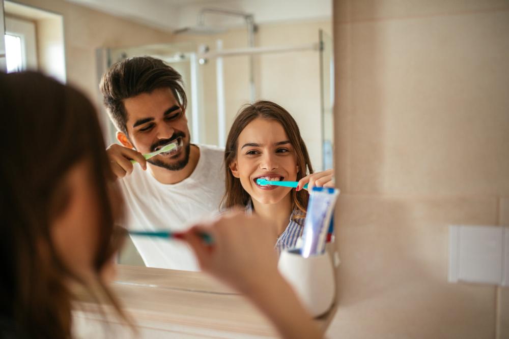 man and woman brushing teeth while looking in mirror.